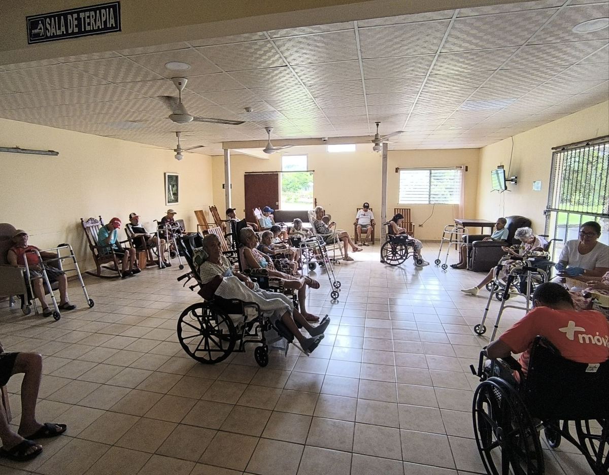 Sala de terapia amplia con residentes en sillas de ruedas y mecedoras, ventiladores de techo y una cuidadora